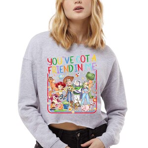 Peut inclure: Un sweat-shirt court gris clair avec une impression graphique color&eacute;e repr&eacute;sentant divers personnages de jouets anim&eacute;s. Au-dessus des personnages, le texte YOU'VE GOT A FRIEND IN ME est &eacute;crit en lettres multicolores. Le motif est bord&eacute; de rouge.