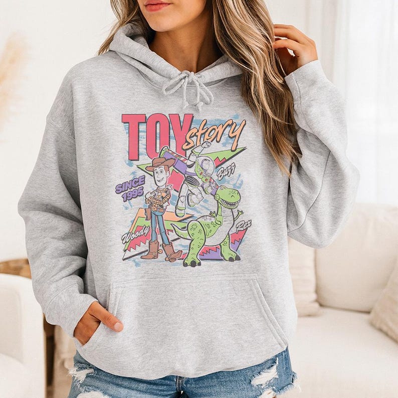May include: A light gray hooded sweatshirt featuring a graphic print of Woody, Buzz Lightyear, and Rex from Toy Story. The design includes the text "TOY story", "SINCE 1995", "Woody", "Buzz", and "Rex" with colorful geometric shapes in the background.