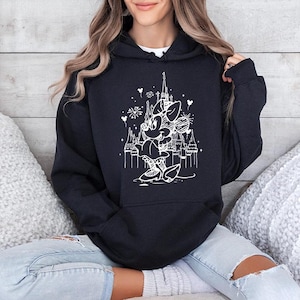 May include: Black hooded sweatshirt featuring a white graphic of Minnie Mouse in front of a castle with fireworks and hearts. The sweatshirt has a drawstring hood and long sleeves. The design is a whimsical illustration.