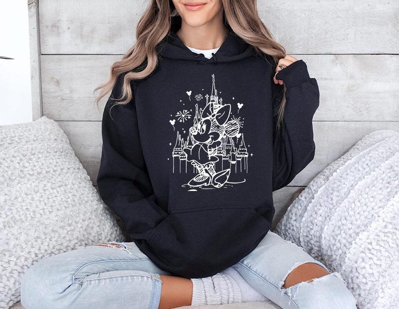 May include: Black hooded sweatshirt featuring a white graphic of Minnie Mouse in front of a castle with fireworks and hearts. The sweatshirt has a drawstring hood and long sleeves. The design is a whimsical illustration.