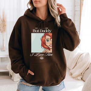 May include: A brown hoodie with a front graphic featuring the text "But Daddy I Love Him" and an illustration of a character with red hair and blue eyes. The hoodie has a drawstring hood and a front pocket.