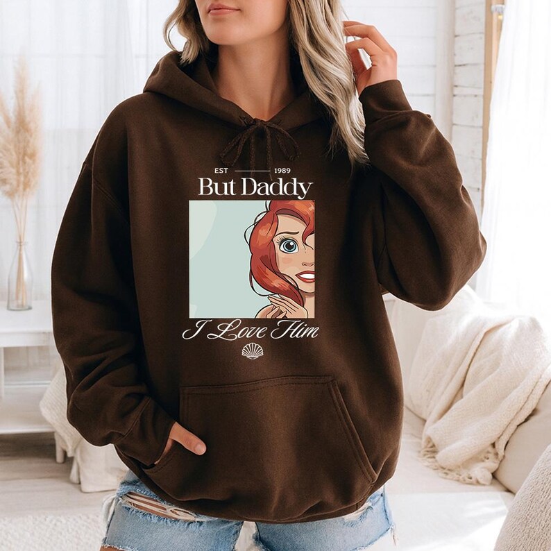 May include: A brown hoodie with a front graphic featuring the text "But Daddy I Love Him" and an illustration of a character with red hair and blue eyes. The hoodie has a drawstring hood and a front pocket.