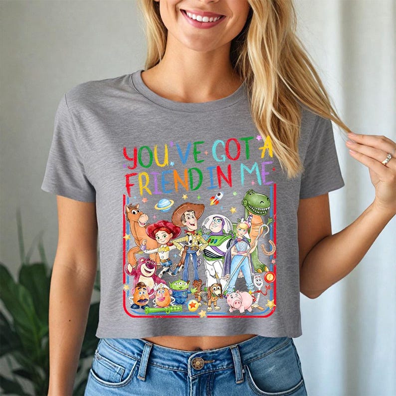 May include: A heather gray short-sleeved t-shirt featuring a colorful graphic of various animated characters from the Toy Story franchise. The text "YOU'VE GOT A FRIEND IN ME" is printed in multicolored letters above the group of characters.