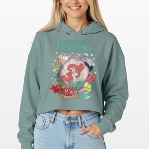 Puede incluir: Una sudadera corta de color verde salvia con un estampado gr&aacute;fico de los personajes de La Sirenita, incluyendo a Ariel en una concha, el Rey Trit&oacute;n, &Uacute;rsula, Flounder y Sebasti&aacute;n, rodeados de burbujas y coral, con el texto The Little Mermaid.
