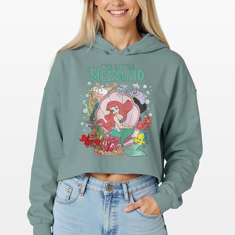 Puede incluir: Una sudadera corta de color verde salvia con un estampado gr&aacute;fico de los personajes de La Sirenita, incluyendo a Ariel en una concha, el Rey Trit&oacute;n, &Uacute;rsula, Flounder y Sebasti&aacute;n, rodeados de burbujas y coral, con el texto The Little Mermaid.