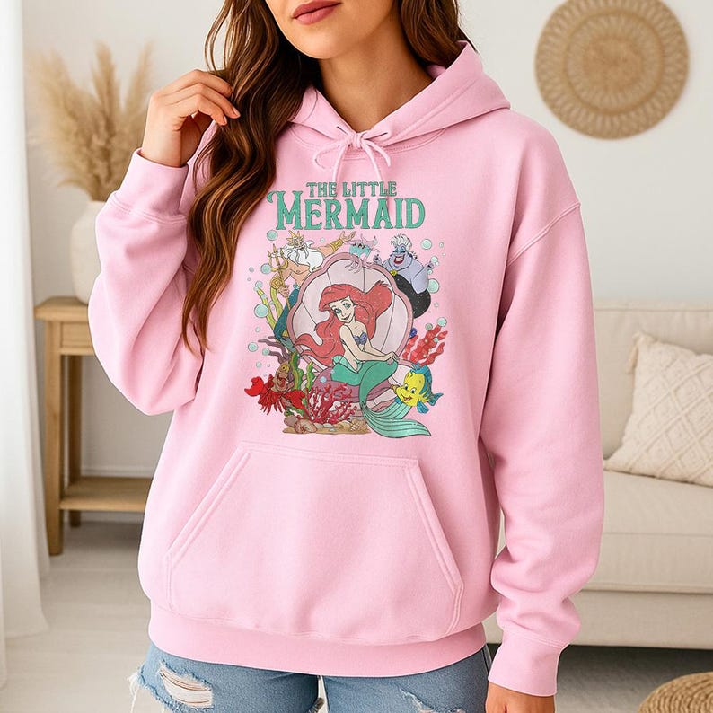 May include: A light pink hooded sweatshirt featuring a graphic print of The Little Mermaid characters including Ariel, King Triton, Ursula, Sebastian, and Flounder. The design includes the text The Little Mermaid in teal lettering above the characters.