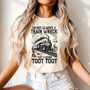 May include: Cream-colored t-shirt with a black and white graphic of a vintage train, cacti, and the text "I'M NOT ALWAYS A TRAIN WRECK JUST KIDDING TOOT TOOT". The shirt has short sleeves and a crew neck.