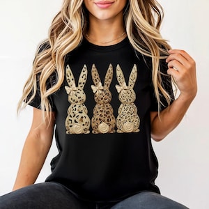 May include: Black t-shirt featuring three decorative bunny figures with leopard print patterns and small bows. The bunnies have a textured, three-dimensional appearance, and are arranged in a row. The design is centered on the shirt.
