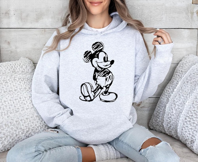 May include: A light gray hoodie featuring a black and white sketch of Mickey Mouse. The hoodie has a drawstring hood and a kangaroo pocket. The design is centered on the front of the sweatshirt.