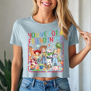 May include: A light blue short-sleeved t-shirt featuring a colorful graphic of various Toy Story characters and the text "YOU'VE GOT A FRIEND IN ME" printed in a multi-colored font. The design includes Woody, Buzz Lightyear, Jessie, and others.