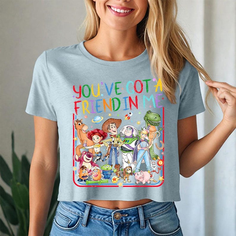 May include: A light blue short-sleeved t-shirt featuring a colorful graphic of various Toy Story characters and the text "YOU'VE GOT A FRIEND IN ME" printed in a multi-colored font. The design includes Woody, Buzz Lightyear, Jessie, and others.
