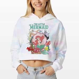 Puede incluir: Una sudadera corta con capucha en tie-dye blanco y azul claro con un estampado gr&aacute;fico de los personajes de La Sirenita, incluyendo a Ariel, el Rey Trit&oacute;n, &Uacute;rsula, Sebasti&aacute;n y Flounder. El texto THE LITTLE MERMAID aparece en verde arriba.