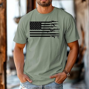 May include: A green t-shirt with a black and white American flag design. The flag has a row of guns across each stripe.