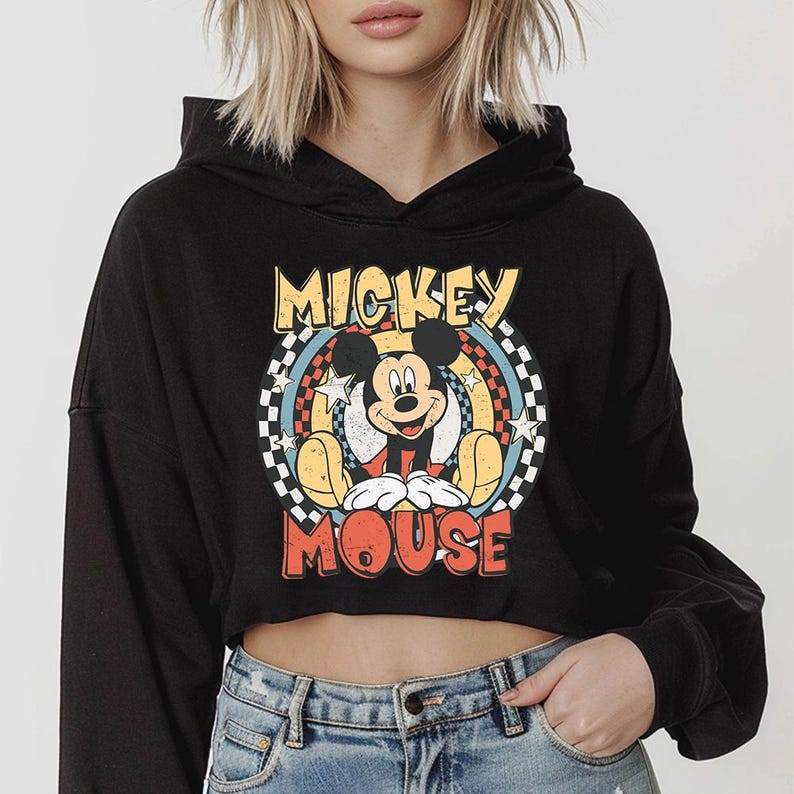 May include: A black cropped hoodie featuring a graphic print of Mickey Mouse. The design includes the text MICKEY MOUSE in yellow and red, surrounded by a circular pattern with a black and white checkered border, blue accents, and white stars.