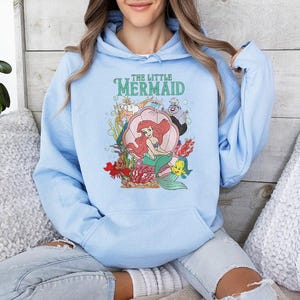 May include: A light blue hooded sweatshirt featuring a graphic print of The Little Mermaid characters, including Ariel sitting in a shell, King Triton, Ursula, and Flounder, with the text The Little Mermaid printed in green above the illustration.