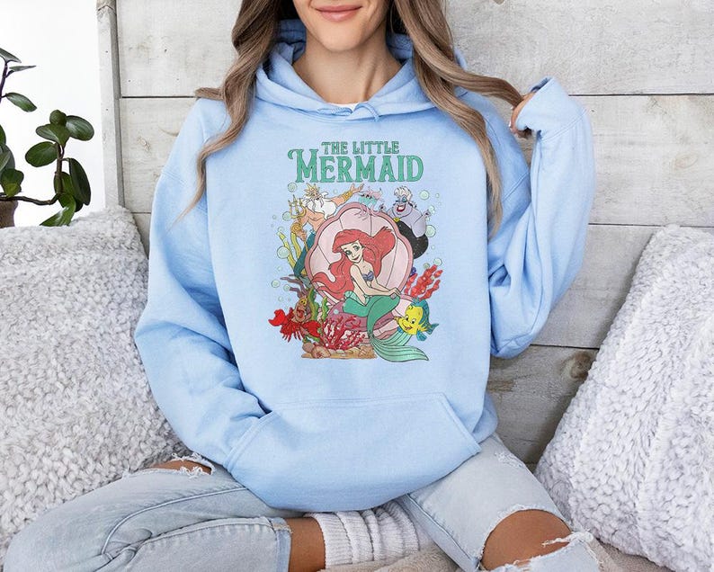 May include: A light blue hooded sweatshirt featuring a graphic print of The Little Mermaid characters, including Ariel sitting in a shell, King Triton, Ursula, and Flounder, with the text The Little Mermaid printed in green above the illustration.