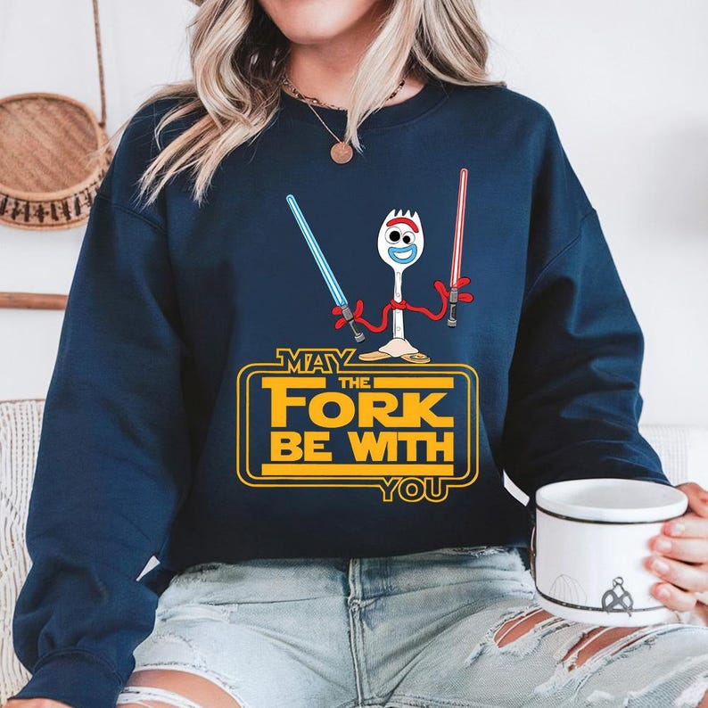 May include: A navy blue long-sleeve sweatshirt featuring a graphic of a white plastic spork character holding a blue light saber in one hand and a red light saber in the other. Below the character is the yellow text May The Fork Be With You.
