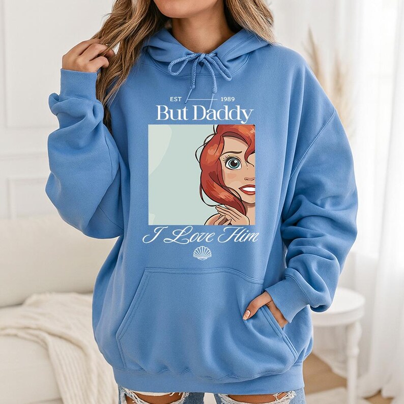 May include: A light blue hoodie with a front graphic featuring a cartoon image of a red-haired character. The graphic includes the text "But Daddy I Love Him" and the year "1989". The hoodie has a drawstring hood and a front pocket.