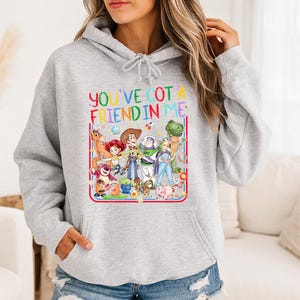 May include: A light gray hooded sweatshirt featuring a colorful graphic print of various animated characters from the Toy Story franchise. The text "YOU'VE GOT A FRIEND IN ME" is printed in multicolored letters above the group of characters.
