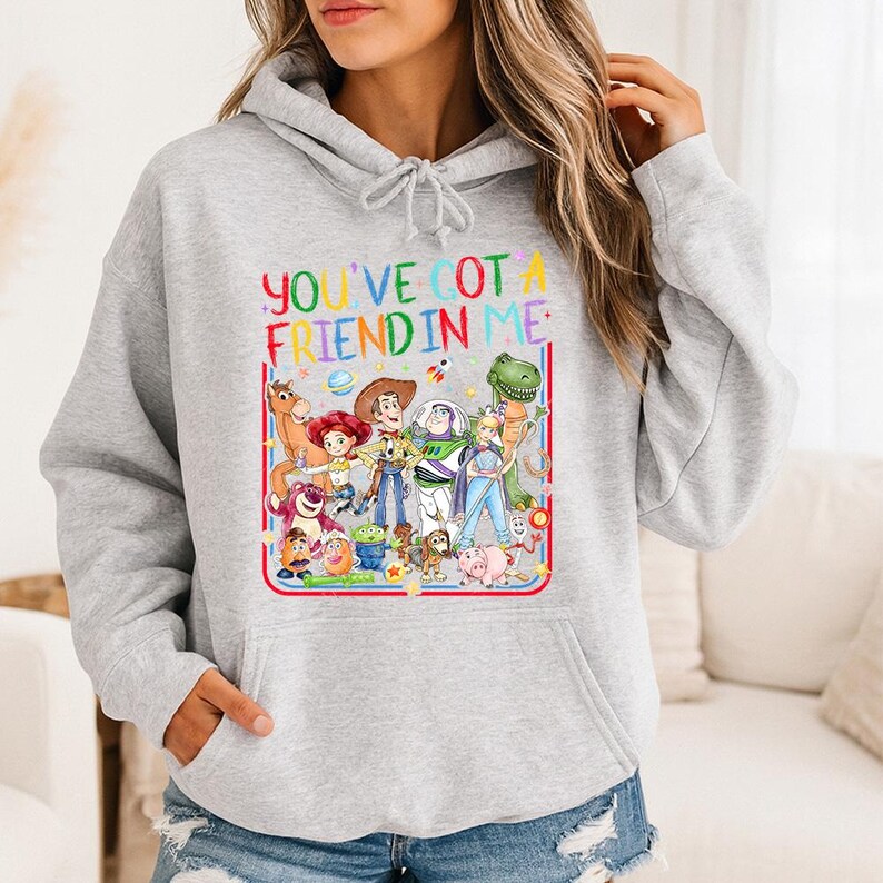 May include: A light gray hooded sweatshirt featuring a colorful graphic print of various animated characters from the Toy Story franchise. The text "YOU'VE GOT A FRIEND IN ME" is printed in multicolored letters above the group of characters.