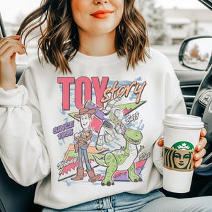 May include: A white crewneck sweatshirt featuring a colorful graphic print of Woody, Buzz Lightyear, and Rex from Toy Story. The design includes the text Toy Story, Since 1995, Woody, Buzz, and Rex, set against a background of geometric shapes.