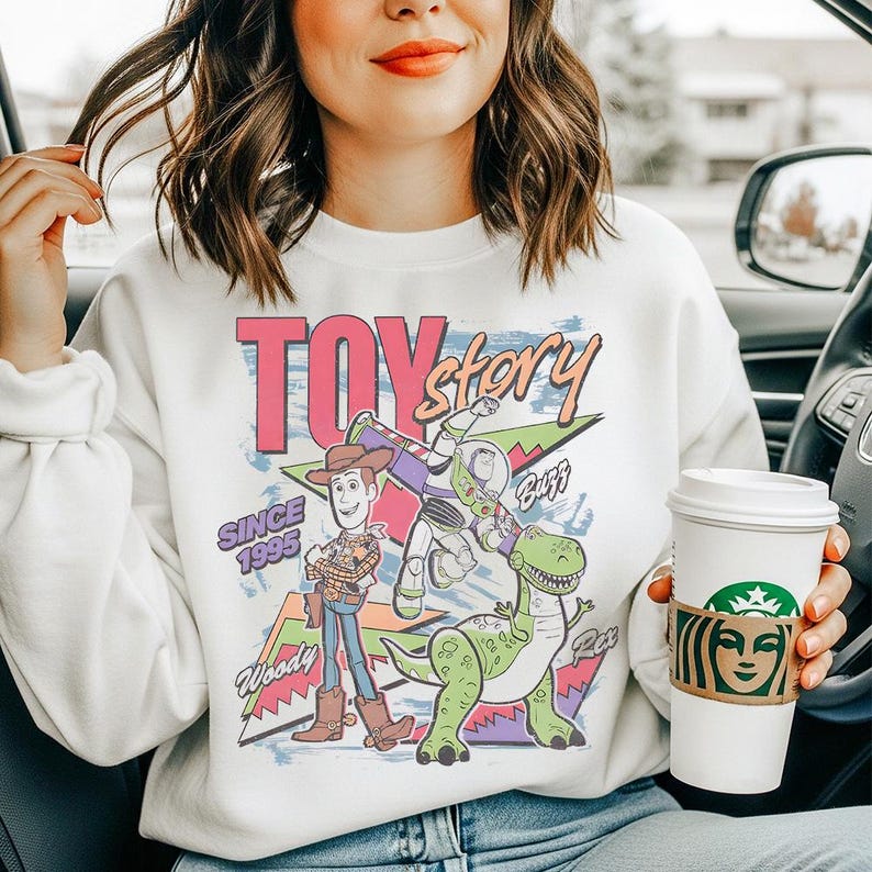 May include: A white crewneck sweatshirt featuring a colorful graphic print of Woody, Buzz Lightyear, and Rex from Toy Story. The design includes the text Toy Story, Since 1995, Woody, Buzz, and Rex, set against a background of geometric shapes.