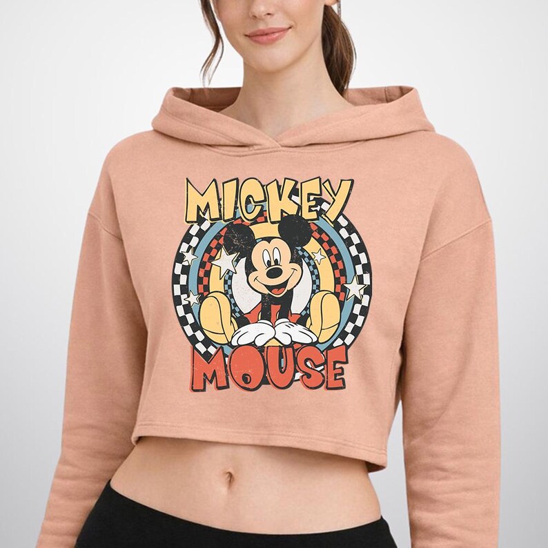 May include: A peach-colored cropped hoodie featuring a graphic print of Mickey Mouse centered within a circular frame. The design includes a black and white checkered border, yellow stars, and the text MICKEY MOUSE in bold, distressed lettering.