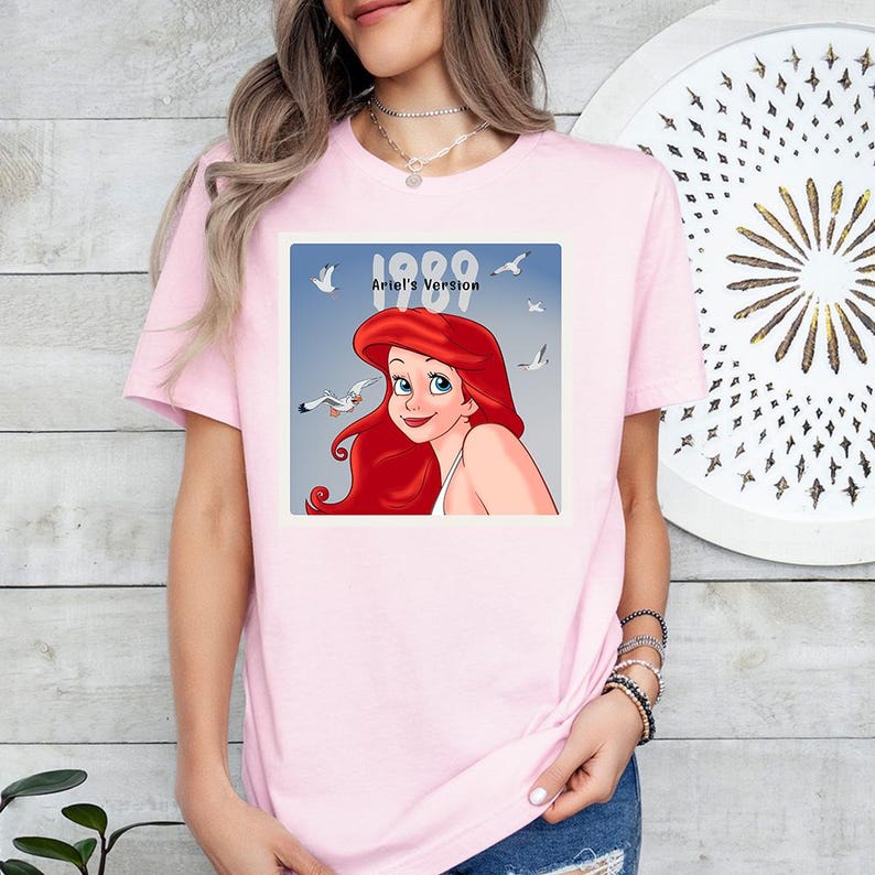 May include: A pale pink t-shirt with a square graphic of Ariel from The Little Mermaid. The graphic shows Ariel with the text "1989 Ariel's Version" and seagulls. The t-shirt has a crew neck.