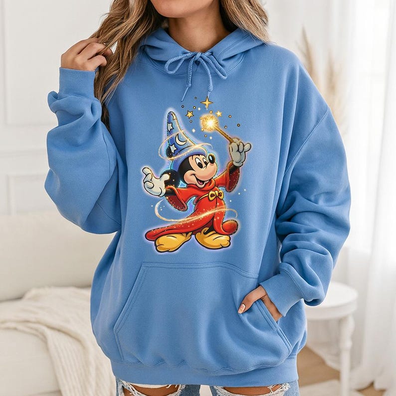 May include: A light blue hoodie featuring a graphic of Mickey Mouse dressed as a wizard, holding a wand with a glowing star. Mickey is wearing a red robe, yellow shoes, and a blue wizard hat. The hoodie has a front pocket and a drawstring hood.