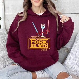 Op de afbeelding: Een bordeauxrode hoodie met een afbeelding van een witte spork die een blauw en een rood lichtzwaard vasthoudt. Onder het figuur staat de gele tekst MAY THE FORK BE WITH YOU, in een rechthoekig kader met afgeronde hoeken.