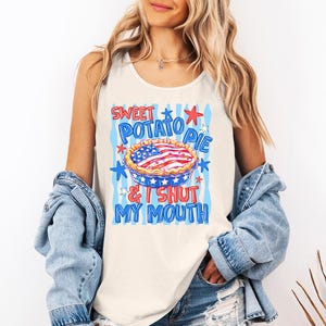 Op de afbeelding: Een crèmekleurige mouwloze tanktop met een grafische print van een taart met een Amerikaans vlagpatroon. De tekst SWEET POTATO PIE & I SHUT MY MOUTH staat in blauwe en rode letters, omringd door blauwe verticale strepen en rode en blauwe sterren.