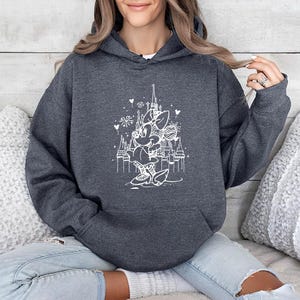 May include: Dark gray hooded sweatshirt featuring a white graphic of a cartoon character in front of a castle. The graphic includes hearts and fireworks. The sweatshirt has a front pocket and a drawstring hood. The model is wearing blue jeans.