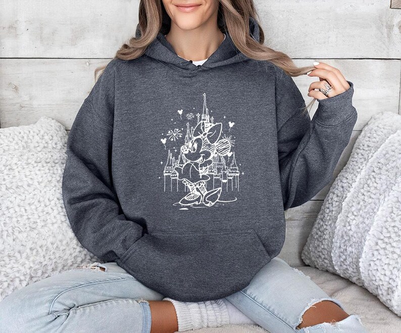 May include: Dark gray hooded sweatshirt featuring a white graphic of a cartoon character in front of a castle. The graphic includes hearts and fireworks. The sweatshirt has a front pocket and a drawstring hood. The model is wearing blue jeans.