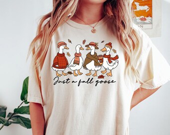 Puede incluir: Camiseta color crema con una ilustración caprichosa de cuatro gansos vestidos con atuendos otoñales. Los gansos llevan suéteres, bufandas y sombreros, uno de ellos sosteniendo una taza de café. El texto "Just a fall goose" está escrito debajo.