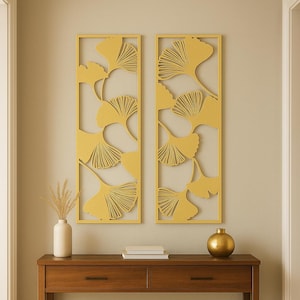 May include: Two rectangular wall art pieces with a yellow ginkgo leaf design hang above a wooden console table. The table has two drawers, a lower shelf, and is topped with a white vase, books, and a gold sphere.