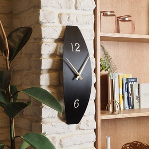 May include: A black oval clock with gold hands and Roman numeral markings. The clock is mounted on a white brick wall.