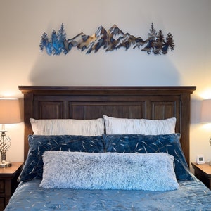 May include: A blue and white patterned bedspread with a fluffy white throw pillow on a bed with a dark wood headboard. A metal mountain range wall art hangs above the bed.