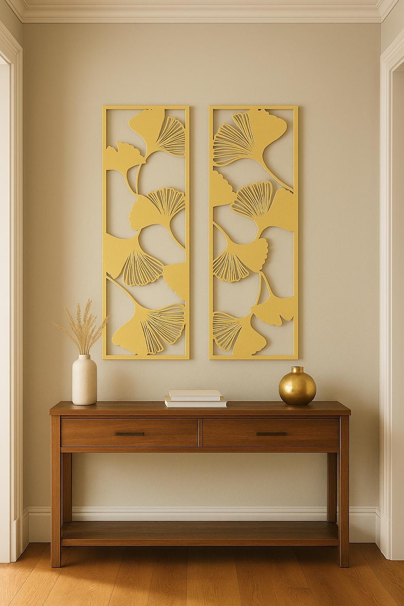 May include: Two rectangular wall art pieces with a yellow ginkgo leaf design hang above a wooden console table. The table has two drawers, a lower shelf, and is topped with a white vase, books, and a gold sphere.