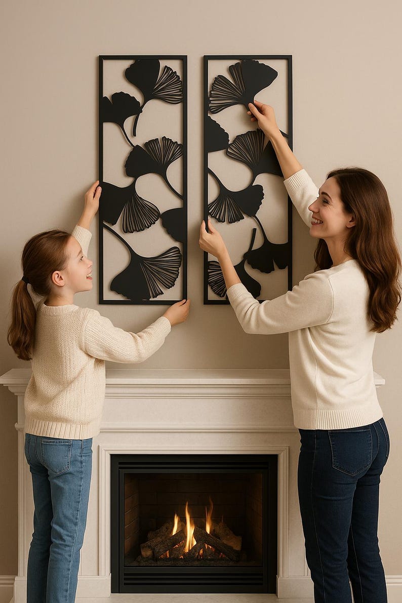 May include: Two black metal wall art pieces with ginkgo leaf designs are being hung on a wall. A woman and a child are shown hanging the art. The art is rectangular and has a black frame.