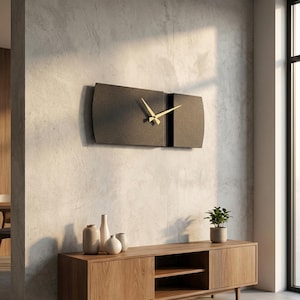 May include: A modern wall clock with a dark gray rectangular face and gold-colored hands. The clock is mounted on a light gray textured wall. The clock's design is minimalist and contemporary, suitable for home decor.