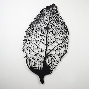 Puede incluir: Escultura de hoja negra estilizada con intrincados cortes en forma de venas. La forma de la hoja es alargada y detallada, mostrando un dise&ntilde;o calado delicado. La obra de arte est&aacute; montada sobre un fondo blanco, resaltando el contraste.