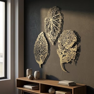 Puede incluir: Tres piezas de arte mural doradas en forma de hoja con intrincados detalles de venas est&aacute;n montadas en una pared gris oscuro. Las hojas son de varios tama&ntilde;os y formas, creando una exhibici&oacute;n art&iacute;stica natural. Debajo hay una consola de madera con objetos decorativos.