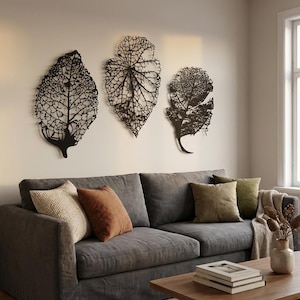 Puede incluir: Tres piezas de arte mural de hojas de metal negro con intrincados detalles de venas est&aacute;n montadas en una pared neutra sobre un sof&aacute; gris. El sof&aacute; est&aacute; adornado con varias almohadas en tonos beige, marr&oacute;n y verde.