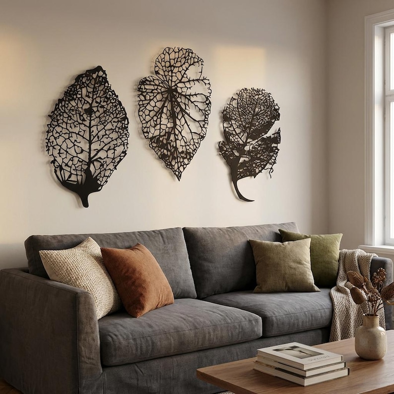 Puede incluir: Tres piezas de arte mural de hojas de metal negro con intrincados detalles de venas est&aacute;n montadas en una pared neutra sobre un sof&aacute; gris. El sof&aacute; est&aacute; adornado con varias almohadas en tonos beige, marr&oacute;n y verde.
