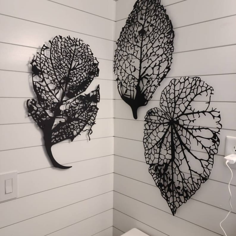 Puede incluir: Tres piezas de arte mural de hojas de metal negro. Cada hoja presenta un dise&ntilde;o intrincado y calado, que muestra las venas y la estructura de una hoja. Las hojas est&aacute;n montadas en una pared blanca con tablones.
