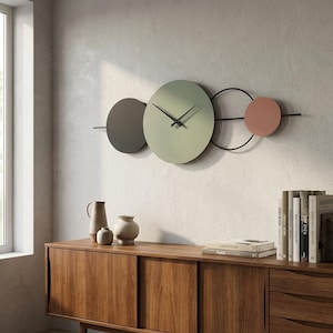 Op de afbeelding: Een moderne wandklok met een minimalistisch ontwerp. Het heeft een lichtgroene cirkelvormige wijzerplaat met zwarte wijzers, geflankeerd door een grijze en een roze cirkel. De klok is gemonteerd op een zwart metalen frame, wat een stijlvolle, eigentijdse uitstraling creëert. De diameter is ongeveer 30 cm.