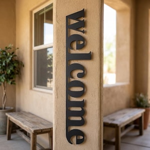 May include: A black metal sign that reads "Welcome" in a vertical orientation. The sign is mounted on a beige pillar, with a window and wooden benches in the background. The sign is a decorative item for a home's entrance.