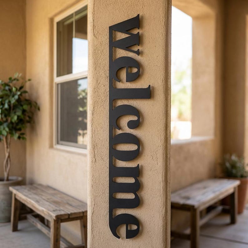 May include: A black metal sign that reads "Welcome" in a vertical orientation. The sign is mounted on a beige pillar, with a window and wooden benches in the background. The sign is a decorative item for a home's entrance.