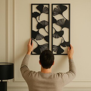 May include: Two black metal wall art panels with a ginkgo leaf design. The panels are rectangular and are being held up against a cream-colored wall. The ginkgo leaves are cut out of the metal, creating a negative space effect.