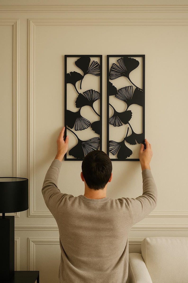 May include: Two black metal wall art panels with a ginkgo leaf design. The panels are rectangular and are being held up against a cream-colored wall. The ginkgo leaves are cut out of the metal, creating a negative space effect.